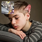 A cozy tabby cat is peacefully napping on a gray cushion, surrounded by a colorful and slightly cluttered background including a pink bra and various household items, creating a casual and homey atmosphere.