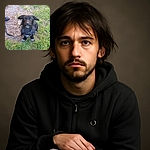 A scruffy black dog with a charmingly tousled coat sits patiently on a patch of grass and gravel, looking up with bright, curious eyes and slightly perked ears. The leash and a sneaker-clad foot hint at a walk in progress, while the natural outdoor setting adds a casual, candid vibe.