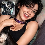 A goofy black dog is flopped on a bed showing a crooked-toothed grin and one ear perked up like it's posing for a silly portrait — flash caught a green-eye glow and the cozy blankets and pastel pillow make it look like royalty on a throne of laundry.
