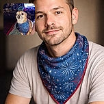 A small dog wearing a festive blue bandana with fireworks print sits attentively on a blanket indoors. The background shows a cluttered room with a TV screen displaying a bright scene, various household items, and a laundry basket. The dog's expression is calm and curious, illuminated by a strong light source.