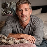 A soulful dog with big, expressive eyes lounges comfortably on a fluffy blanket, surrounded by cozy bedding and pillows. The lighting is soft, highlighting the dog's gentle expression, while the text overlay lovingly declares 'My boy Blue' with a blue heart emoji.