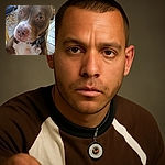 A close-up photo of a brown and white dog with soulful eyes looking directly at the camera, standing on a wooden porch with a car and some leaves visible in the background.
