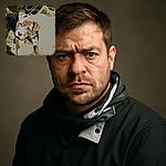 A curious brindle dog dressed in a tuxedo jacket tilts its head inquisitively, standing outdoors on grass with some blurred chairs in the background. The lighting is a bit harsh, causing some overexposure on parts of the dog and ground.