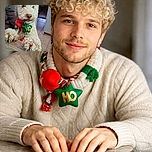 A fluffy white dog with curly fur is standing with its front paws on a table, holding a festive green star-shaped toy with 'HO' written on it in its mouth. The background shows a cozy indoor setting with a tiled floor and some furniture, adding a warm homey vibe.