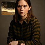 A cozy tabby cat nestled on a soft cream cushion under a wooden structure, peering out with curious eyes in a warmly lit room with wooden flooring and a carpet edge visible.