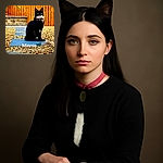 A sleek black and white cat named Minnie sits majestically on a blue stepping stone surrounded by a bed of pebbles, with a warm wooden fence and a small palm tree in the background. The cat's green eyes are striking against its dark fur, and it wears a collar with a bell and tag. The photo has a bright and vibrant outdoor setting with a clear focus on the poised feline.