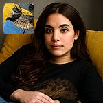 A fluffy tabby cat lounges comfortably on a couch adorned with colorful cushions, looking curiously at the camera with wide, bright eyes that sparkle like tiny emeralds.