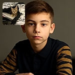A curious tabby kitten is cozily nestled inside an open black bag on a patterned bedspread, looking straight at the camera with wide, innocent eyes as if ready to pounce on any unsuspecting laptop or homework.