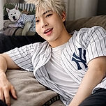 A fluffy white dog lounging comfortably on a bed, wearing a striped shirt that adds a touch of sporty charm. The background is a cozy mix of pillows and blankets, making the dog look like it's ready for a relaxed day in or maybe a playful nap session.