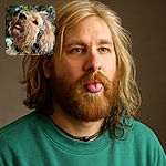 A close-up shot of a wet, scruffy dog with its tongue slightly out, looking happy and playful in a sunlit forest setting. The dog's curly fur is a mix of cream and black, and there's a tattooed arm in the background, suggesting a loving owner nearby.