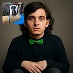 A curious black puppy with a green tag sits atop a large, taped-up cardboard box in a cluttered garage or workshop filled with tires, tools, and various equipment, looking directly at the camera with innocent eyes.