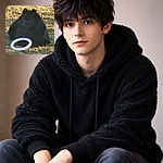 A fluffy black dog lounges in the grass like a tiny, suspicious shadow, guarding its white frisbee ring and pondering whether to chase or nap — dramatic ears, soulful eyes, and maximum fluffiness.