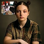 A close-up photo of a black and white Boston Terrier dog lounging on a colorful plaid blanket, looking directly at the camera with a curious expression, one eye slightly red.
