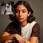 A close-up photo of a brindle and white dog lying on a bed covered with light-colored blankets, gazing curiously at the camera with soulful eyes in a dimly lit room.