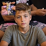 A wide-eyed tabby cat sits proudly in someone’s lap, wearing a shiny gold chain and a tiny tag like it just became the neighborhood’s most serious celebrity. Behind it, a person holds a phone while colorful clothing and a dark indoor background complete the candid portrait.
