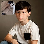 A curious black and white puppy stands on a tiled floor looking back towards the camera, next to a person's foot clad in a black sandal and white sock, with a wooden kitchen cabinet in the background.