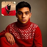 A cute black cat wearing a festive red and white sweater looks up with curious eyes, sitting on a wooden floor between a person's red pants. The cozy indoor setting and the cat's shiny fur make for a charming and warm snapshot.