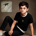 A slightly blurry photo of a black and white cat lounging on a wooden floor, looking curiously at the camera with a collar tag shining.