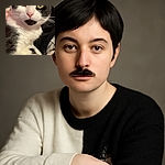 A close-up photo of a black and white cat with striking yellow eyes and a distinctive black patch on its chin, looking directly at the camera. In the bottom left corner, a small video call window shows a man holding a dark object, possibly a cat toy or another pet. The background features a warm reddish wall and some household items.