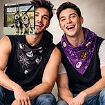 Two happy huskies wearing festive Halloween bandanas pose in front of a chain-link fence adorned with colorful locks, with a beautiful sunset reflecting off the water behind them. Their smiles and the glowing sky create a warm, joyful scene.