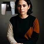 A calico cat sits attentively on a dark rug between a light wooden table and a cozy couch draped with textured blankets, looking directly at the camera with curious eyes.