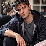 A cozy gray cat with a shiny green bell collar is lounging comfortably on a soft pink blanket, surrounded by a homey background of bookshelves and a black chair, looking slightly sleepy or contemplative.