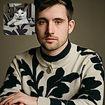 A poised and regal cat lounges elegantly on a patterned couch, staring directly into the camera with calm, inquisitive eyes. The background features a softly blurred lamp and subtle home decor, giving the scene a cozy and intimate vibe.
