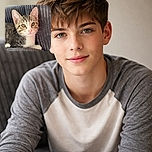 A charming tabby kitten with bright eyes and a pink nose lounges comfortably on a textured gray blanket and cushion, looking curiously at the camera with soft natural lighting highlighting its fur.
