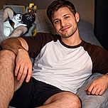 A contented dog sprawled across a man's lap like the boss of the sofa, giving a goofy grin to the camera while the owner lounges behind with a relaxed, phone-in-hand look. Flash highlights whiskers and a collar, the couch's quilted back provides a cozy backdrop, and the whole scene reads "prime weekend nap time".