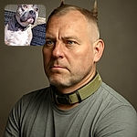 A close-up photo of a muscular dog with a focused expression, sitting on a woven outdoor chair with textured wall background, basking in warm sunlight that highlights its fur and eyes.