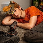 A cozy orange cat is napping peacefully on a gray carpet, nestled beside a cordless drill and some tools, while a boot lies nearby. The background shows a shelf with quirky items including a green dragon figurine, a pineapple, a Rubik's cube, and some books, adding a touch of whimsy to this handyman's resting feline moment.