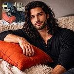 A fluffy black and brown dog lounges regally on a fluffy beige blanket, resting its paw on a bright orange cushion like a royal on a throne in a cozy indoor setting.