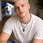 A curious white dog with a black collar sits on a carpeted floor, tilting its head slightly as if trying to understand the camera, with a dark leather couch and some household items in the background.