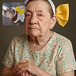 A small dog dressed in a colorful floral outfit with a large yellow bow and a white lace headband looks up with wide, curious eyes, standing on a carpeted floor with sunlight casting shadows nearby.
