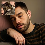 A close-up of a cozy tabby cat lying on a bed with a pink pillow in the background, looking relaxed with its tongue slightly sticking out, while a hand gently pets it.