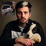 A cozy black and white dog lounges comfortably in a plush pet bed, resting its head on a soft stuffed duck toy, surrounded by other scattered toys on a carpeted floor in a warmly lit room.