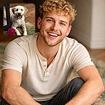 A cheerful fluffy dog with curly cream and tan fur is lying on a multicolored rug next to a bright pink spiky ball, tongue out in a happy pant, indoors on a wooden floor with a piece of furniture nearby.