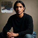 A sleek black dog lounges on a pile of blankets on a tiled floor, with a chew toy nearby and a vintage-looking black dresser in the background. The dog looks relaxed but alert, blending a bit into the dim lighting and dark furniture.