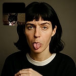 A grainy, low-light photo of a black and white cat sticking out its tongue in a silly manner, with a dark and cluttered background.