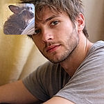 A scruffy little dog giving an earnest 'who, me?' expression — one big ear perked, shiny curious eyes, and a tousled beard of fur. The pup is snuggled against a soft couch with a warm curtain/wall background, looking up as if auditioning for a tiny dramatic role.