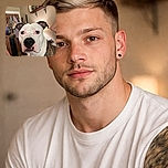 A close-up photo of a serious-looking brindle and white dog with a black nose, sitting indoors with a cozy background of a door, hanging clothes, and a shelf with framed pictures. The dog seems to be the main focus, gazing intently at the camera.