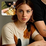 A curious calico cat with bright green eyes gazes upward, nestled comfortably against a person's arm and torso. The person wears a white shirt and black pants, with a tattoo visible on their leg. The background features a soft green surface, creating a cozy and intimate atmosphere.