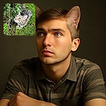 A curious tabby cat with striking blue-green eyes is crouched in the grass near a black chain-link fence, looking intently upwards as if spotting a secret squirrel or a flying treat.