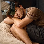 A cozy brown dog is curled up napping on a fluffy white rug on a wooden floor, surrounded by a few household items including a bag, a pet stair, and a headset hanging nearby.