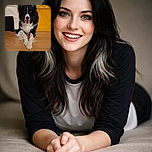 A happy black and white dog is lying on a wooden floor with its front paws crossed, looking straight at the camera with a joyful expression. Behind it, a cream-colored couch and a bright yellow textured cushion add a cozy and colorful background.