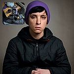 A curious black dog with a purple cloth wrapped around its head peeks out from a black backpack, sitting in a cluttered room with a couch, kitchen utensils, and a wire crate in the background.