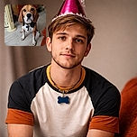 A beagle wearing a shiny pink party hat sits obediently on a wooden floor, staring intently at the camera. In front of the dog, a plate holds a sausage shaped like a smiley face decorated with green bits. The background shows a wooden dresser, a fan, and some household items, creating a cozy indoor scene.