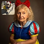 A fluffy small dog dressed in a colorful Snow White-inspired costume with a red bow sits adorably on a cushioned wicker chair outdoors, basking in the sunlight on a patterned stone patio.
