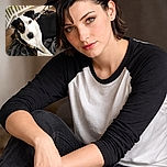 A charming dog with a striking black and white coat lounges comfortably on a textured gray blanket, looking curiously at the camera with warm amber eyes. The background is softly blurred, highlighting the dog's expressive face and relaxed pose on a wooden floor near a white railing.