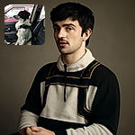 A well-focused photo of a black and white dog sitting patiently in the passenger seat of a car, wearing a harness. The background shows a red car parked outside, and the overall lighting is natural with good detail on the dog's fur.
